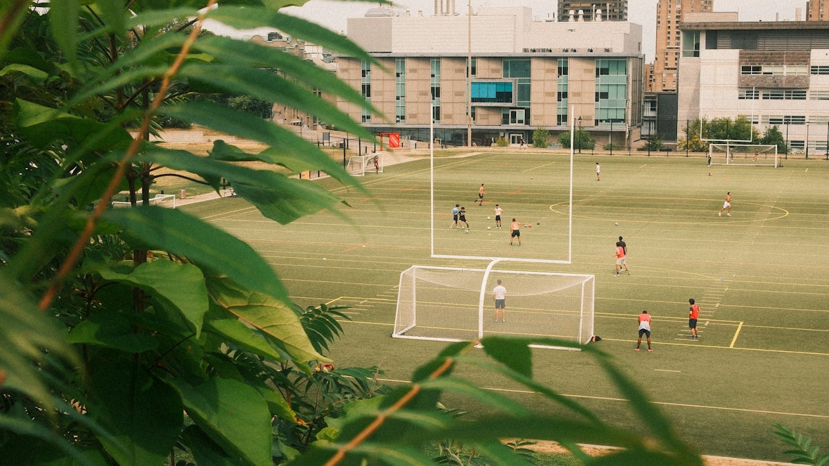 People playing soccer on a field with buildings behind.